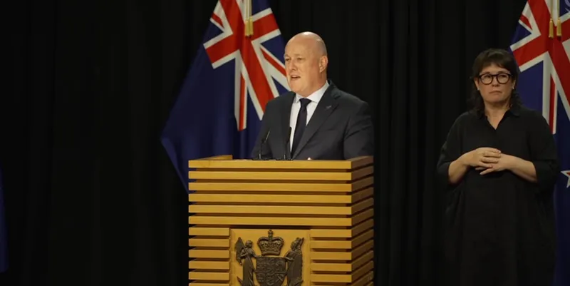 A man in a suit speaks at a wooden podium with the New Zealand coat of arms, with a New Zealand flag behind him; a woman stands beside him signing in New Zealand Sign Language.