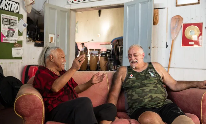Two older men sit on a pink couch, laughing and talking in a cozy, decorated room with posters, an oar, and drums in the background. One wears a red shirt, the other a green tank top and shorts.