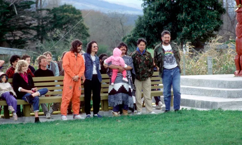 A group of people, including a child in pink, stand and sit on benches outdoors near a carved wooden figure, surrounded by greenery and hills in the background.