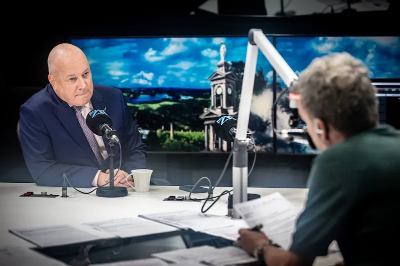 A bald man in a suit sits at a radio studio desk with a microphone, speaking to another person holding papers. A large screen behind them displays an outdoor scene with a statue and greenery.