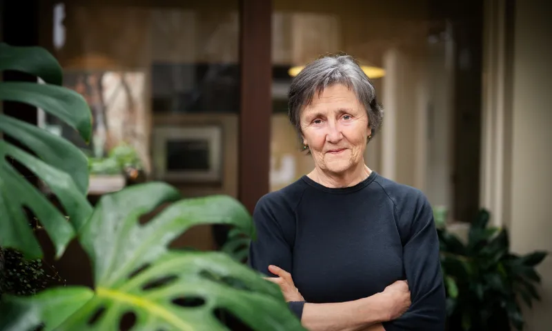 A photograph of an older woman with short grey hair: she is in the background with a plant in the fore. She is wearing a black t-shirt, is folding her arms and is staring at the camera.