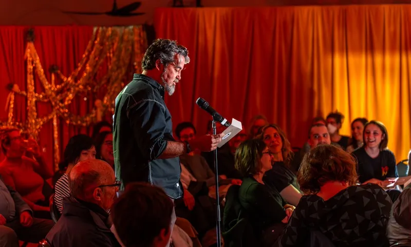 A man stand in front of a microphone and reads from aa card he is holding in his hand. Behind him are the theatre audience