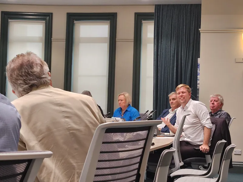 Several people sit around a conference table in discussion. One man in a white shirt is speaking and gesturing with his hand, while others listen. The room has large windows and office chairs.