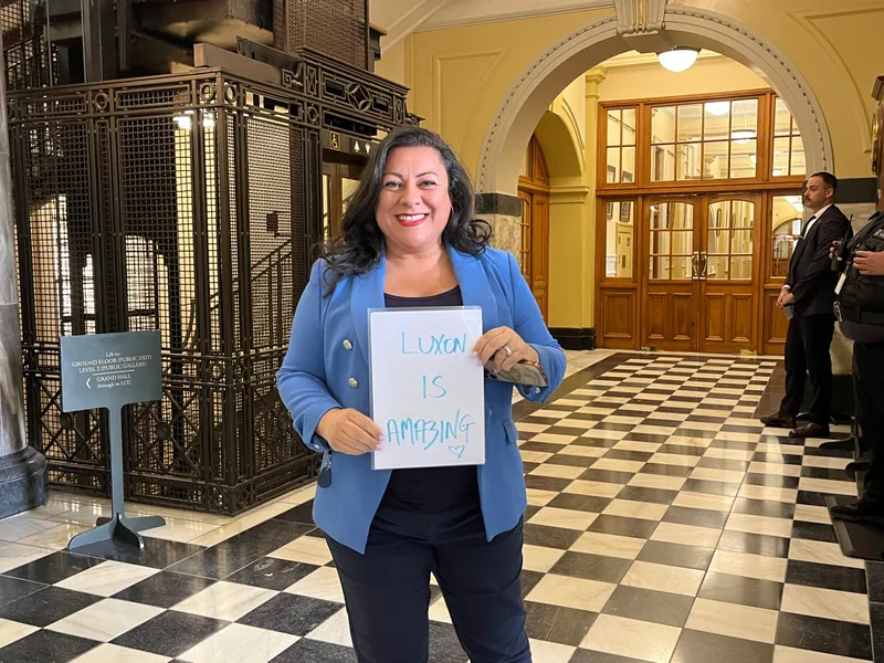 A smiling woman in a blue blazer holds a sign reading "Luxon is amazing" while standing in a hallway with black-and-white checkered floors and ornate decor. Two people stand in the background near a doorway.
