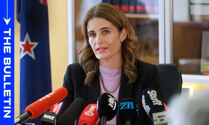 A woman with wavy brown hair sits at a desk, speaking at a press conference surrounded by microphones. The New Zealand flag is visible behind her, along with shelves of books. A blue graphic reads "The Bulletin.