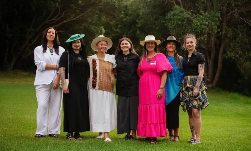 Seven women stand together outdoors on green grass, smiling. They wear a mix of colourful and neutral outfits, including dresses, hats, and accessories, with trees and bushes in the background.