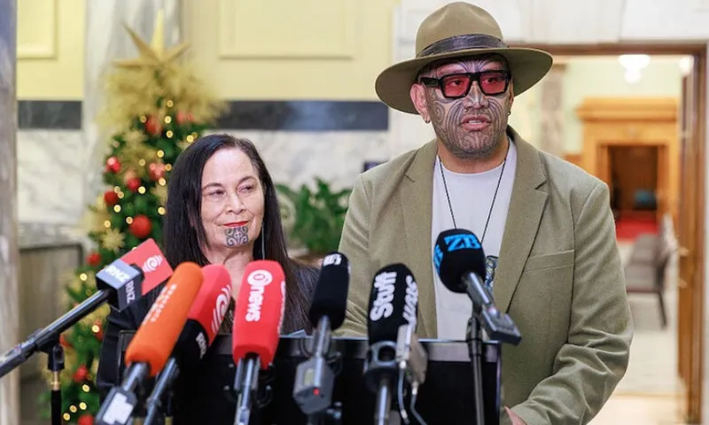 Two people with traditional Māori chin tattoos stand at a podium surrounded by microphones, giving a press conference in an indoor setting with a Christmas tree visible in the background.