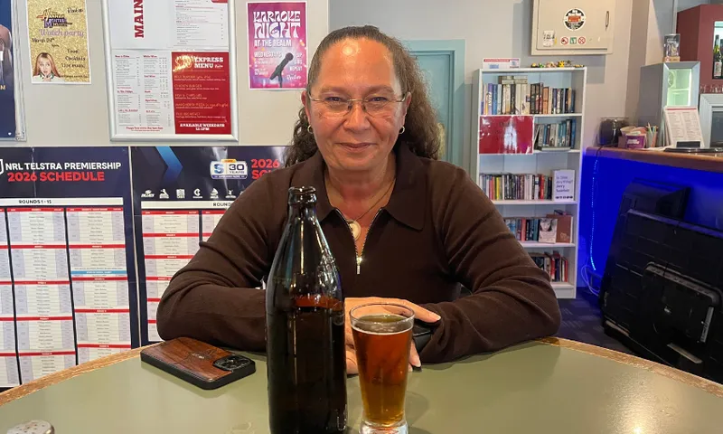 a woman with a slicked back ponytail and glasses sits at a high bar table with a brown beer bottle and pint of beer in front of her