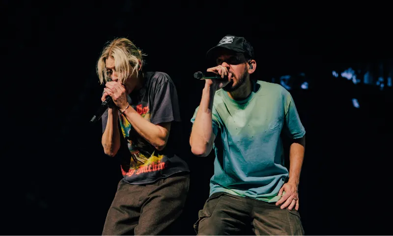 Two musicians performing on stage, both holding microphones. One wears a graphic t-shirt and has blond hair, while the other wears a cap and a plain t-shirt. The stage background is dark.