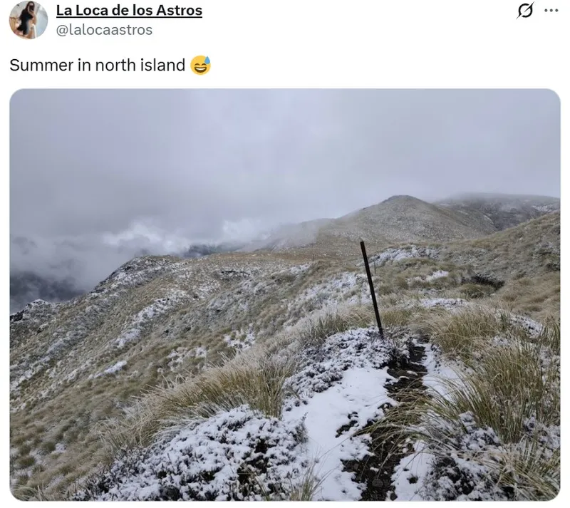 A mountain landscape on a cloudy day with light snow covering grassy slopes and a narrow trail marked by a pole. The scene looks cold and wintry despite being summer.