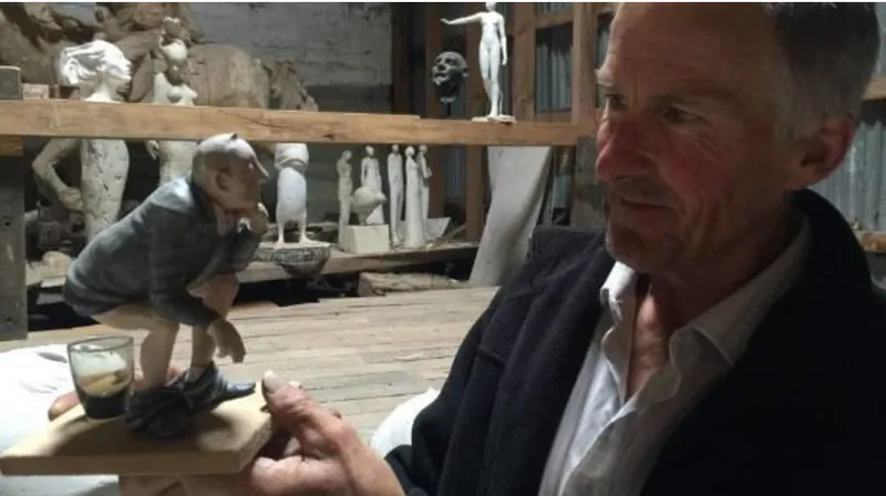A photo of an elderly man with a miniature sculpture of a man squatting, pants down, over a glass of water. There are other sculptures in the background, he is clearly an artist.