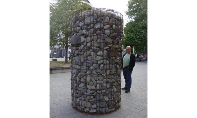 A photo of a stone cairn: a pile of riverstones contained within a wire frame and taller than a tall person.