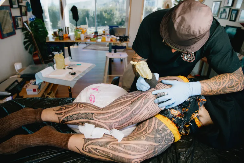 Tattoo artist working on a client’s leg while the client lies on a table, showing extensive patterned tattoo coverage across both legs.