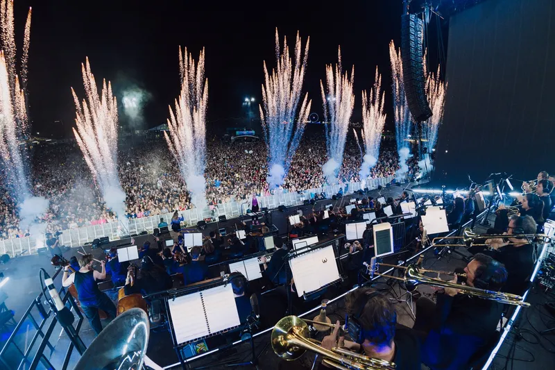 A large orchestra performs on stage before a packed outdoor audience at night, with fireworks shooting into the sky and bright stage lights illuminating the scene.