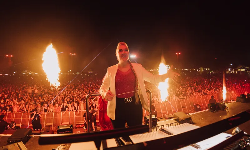 A conductor in a white jacket leads an orchestra onstage at night, with large flames shooting up beside her and a huge crowd cheering in the background. The scene is energetic and dramatic.