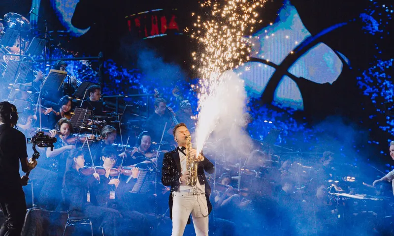 A performer in a sparkly jacket releases fireworks from a handheld device on stage, with an orchestra playing behind him and blue, abstract visuals on a large screen in the background.