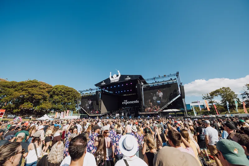 A large crowd gathers outdoors in front of a stage with big screens at a music festival on a sunny day. The stage displays "The Exponents," and people are enjoying the live performance.
