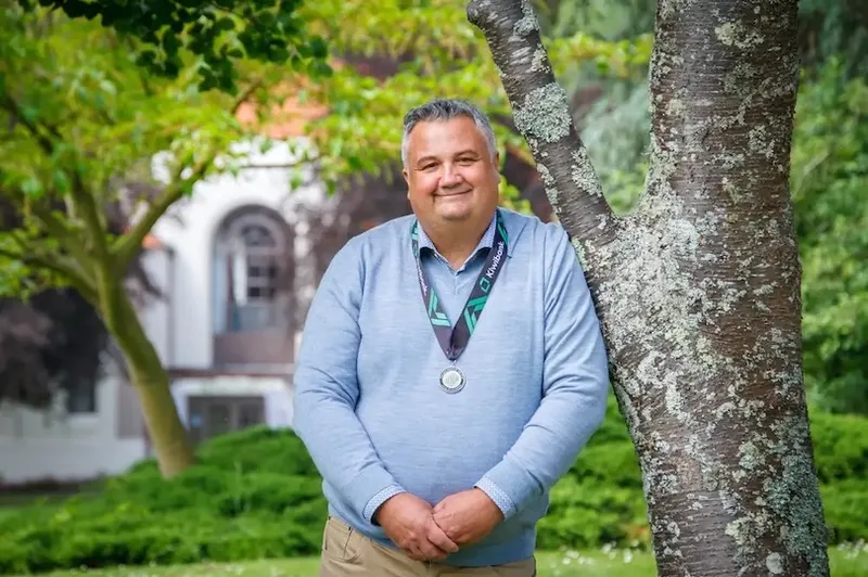 A man wearing a light blue sweater and a medal on a ribbon stands outdoors, leaning against a tree with green foliage. A building with arched windows is visible in the blurred background.