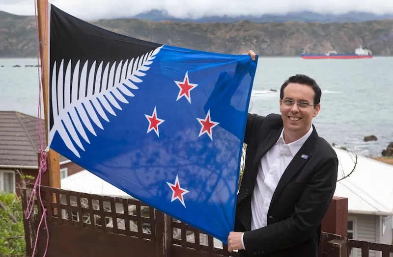 A person in a suit smiles while holding a New Zealand flag with a silver fern design, standing by a wooden fence near the water with hills and a ship in the background.