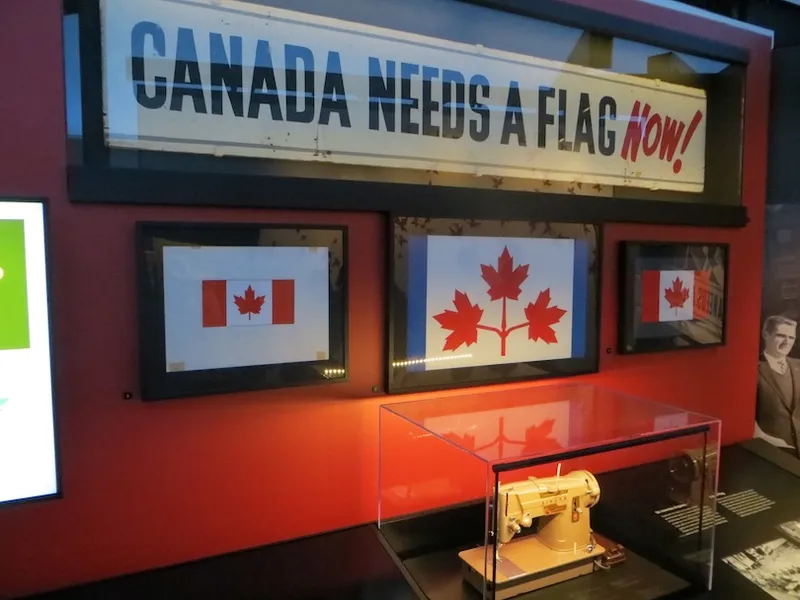 A museum display shows two framed Canadian flag designs and a sewing machine under glass, with a large vintage sign above reading "CANADA NEEDS A FLAG NOW!" in bold letters.