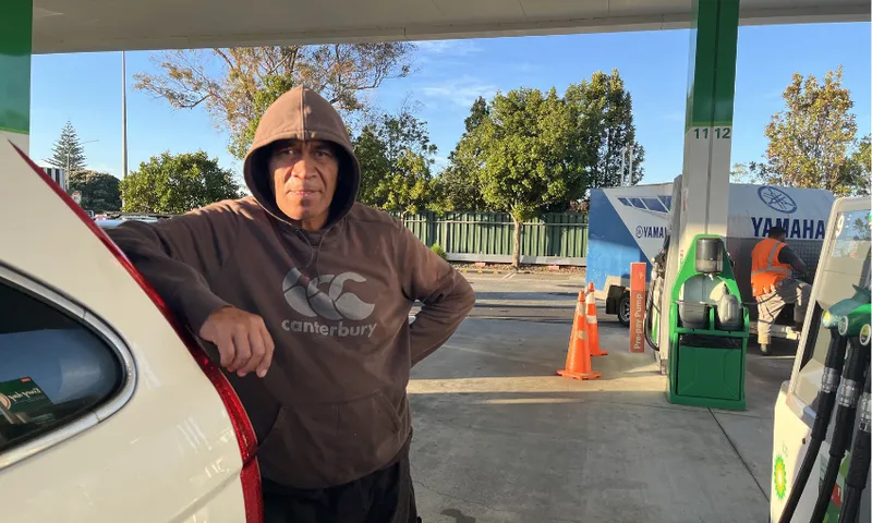 A man in a brown hoodie stands next to a white car at a gas station during the day. He is leaning on the car, with one hand on his hip. Fuel pumps and orange cones are visible in the background.