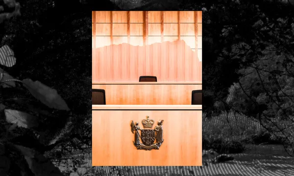 A wooden judge’s bench with an empty chair and a coat of arms emblem is seen in a courtroom, set against a backdrop of wooden paneling and windows.