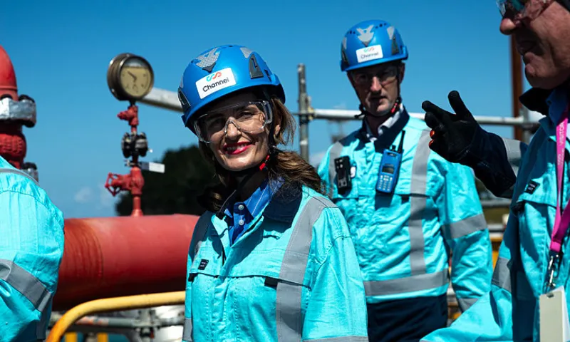 A group of people in blue safety gear and helmets stand outdoors near industrial piping and gauges. A woman in front is smiling at the camera, while others are engaged in conversation.