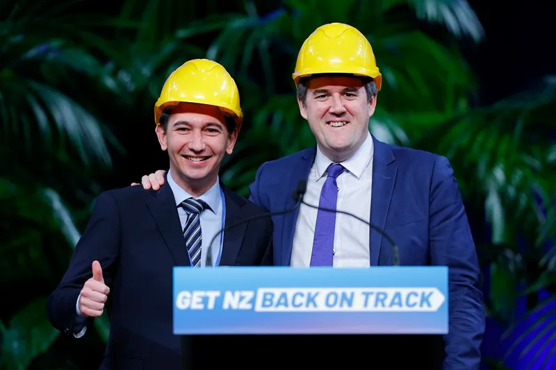 Two men in suits wearing yellow construction helmets stand smiling behind a podium that reads “GET NZ BACK ON TRACK.” One gives a thumbs-up; green plants are visible in the background.