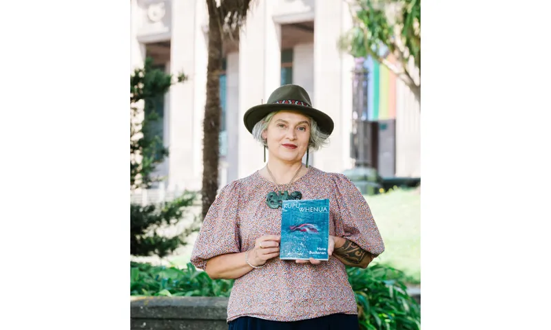 A photo of Hana Buchanan, who is standing outside holding up a copy of her book. She's wearing a hat and is smiling.