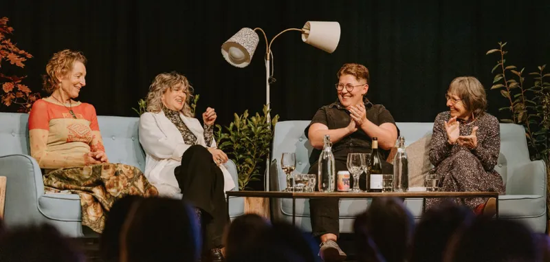 Four women are sitting on a two couches on a stage and are all laughing and smiling in front of a live audience.