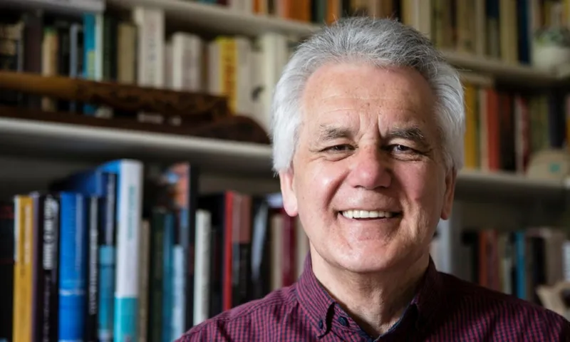 An older man with white hair and a red checkered shirt smiles in front of bookshelves filled with colorful books.