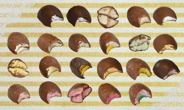 Rows of chocolate-covered sweets, each cut in half to show colorful fillings in shades of white, yellow, green, pink, and brown, arranged neatly on a striped yellow and white background.