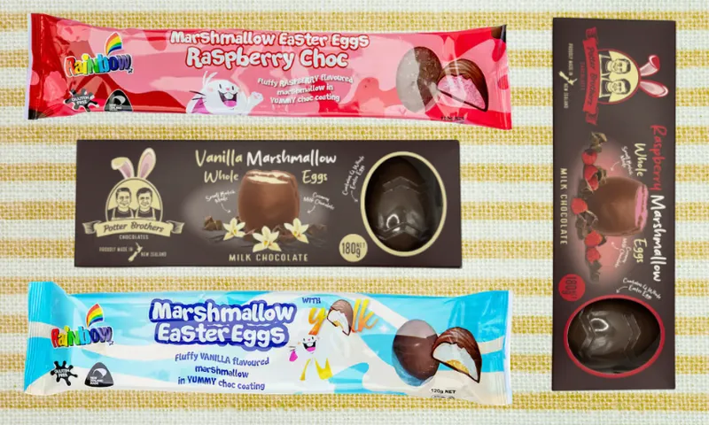 Four different packages of marshmallow Easter eggs, including raspberry choc, vanilla marshmallow, and marshmallow with jelly, displayed on a light striped surface. The packaging features colorful designs and chocolate egg images.