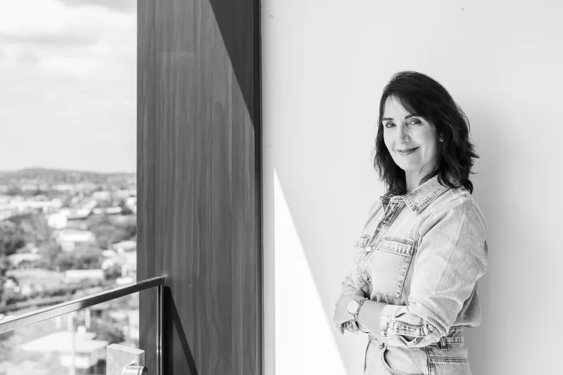 A woman with shoulder-length dark hair stands indoors by a large window, smiling with her arms crossed. She wears a light denim shirt and jeans. Outside, a blurred cityscape is visible. The image is black and white.