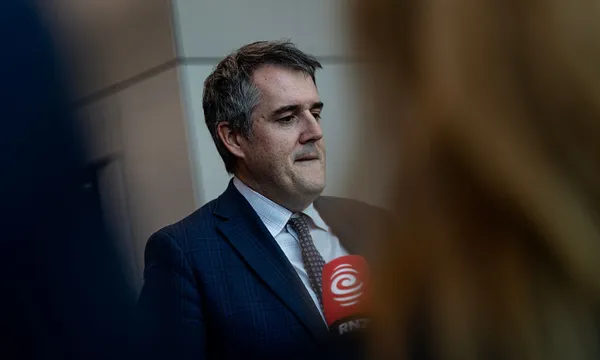 A man in a suit and tie stands indoors, holding a red RNZ microphone. He appears to be speaking or preparing to speak, with blurred figures and a wall in the background.