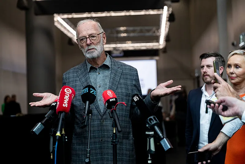 An older man with gray hair and glasses stands at a podium surrounded by microphones, shrugging his shoulders. Two people stand behind him, and reporters hold out recording devices in a modern indoor setting.