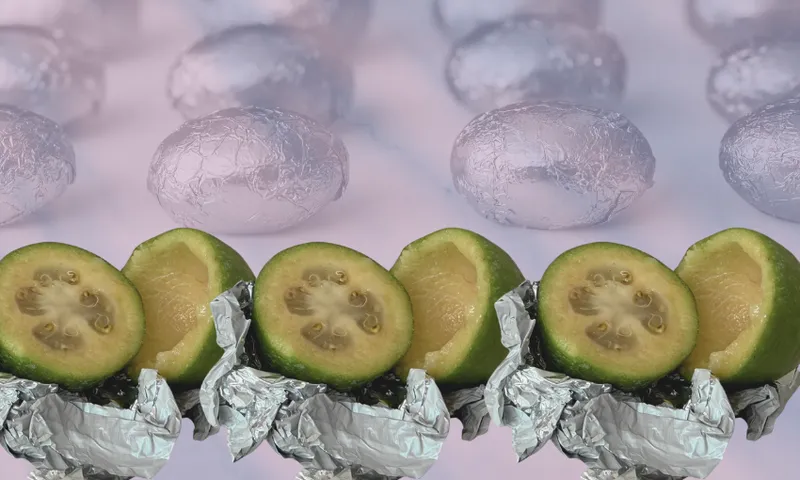 Three halved feijoas wrapped in crumpled aluminium foil are arranged in a row, with a background of silver, foil-wrapped oval objects on a soft purple surface.