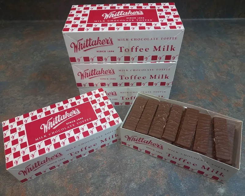 Four boxes of Whittaker’s Toffee Milk milk chocolate toffee are stacked, with one box open showing rows of individually wrapped chocolate pieces inside. The boxes are white with red and silver accents.