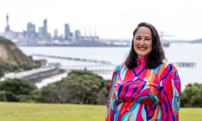 A woman in a colorful, patterned dress stands smiling outdoors with a city skyline, water, and greenery in the background.