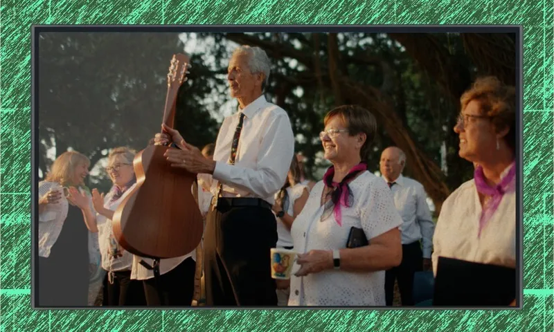 A group of older adults, some holding musical instruments and mugs, stand outdoors under trees. They wear white shirts, black pants, and purple scarves, smiling and appearing ready to perform.