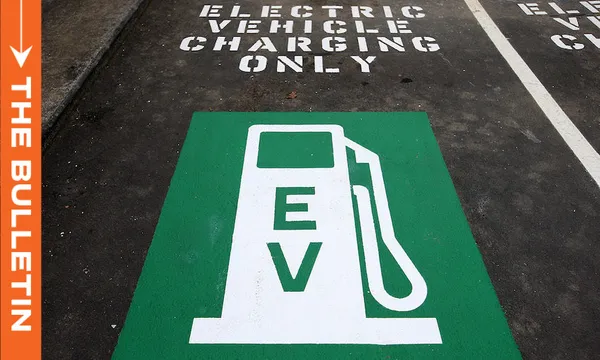 A green pavement marking with a white electric vehicle (EV) charging symbol and the letters "EV" is shown in a parking space labeled "Electric Vehicle Charging Only." An orange banner on the left reads "THE BULLETIN.