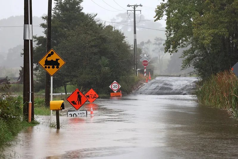 a semi-rural intersection is flooded with muddy rainwater following a cyclone. A number of orange temproray road signs stick out of the water