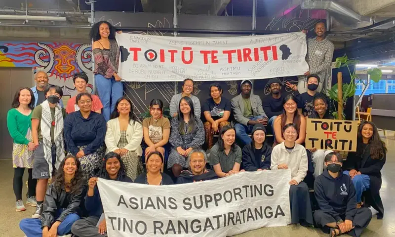 A diverse group of people pose indoors holding signs that read "TOITŪ TE TIRITI" and "ASIANS SUPPORTING TINO RANGATIRATANGA." They are smiling, and some are seated while others stand behind them.