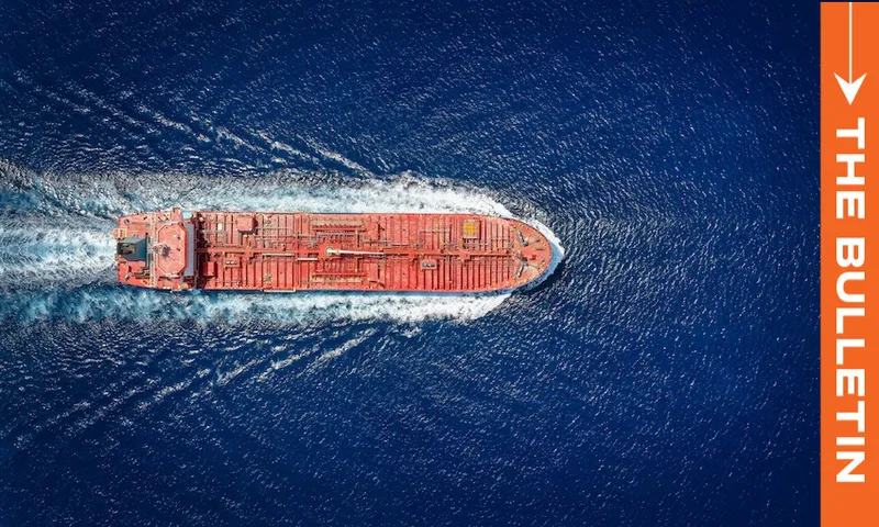 Aerial view of a large red cargo ship moving through deep blue ocean water, creating a white wake. An orange vertical banner on the right reads "THE BULLETIN" with a white arrow above the text.