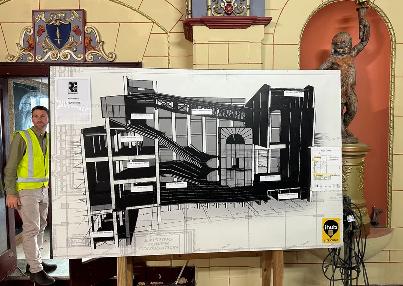 A large display showing black and white plans for a theatre. in the back is a tiled wall and a man wearing a high vis vest