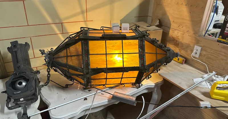 A very old cast iron lamp with black framing and a warm light glowing within sits on an old white painted table