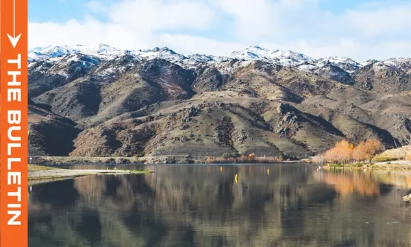 A calm lake with clear reflections sits in front of rugged, snow-dusted mountains under a partly cloudy sky. On the left, an orange vertical banner reads "THE BULLETIN" in white text.