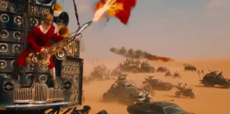 A man in a red outfit plays a guitar that shoots flames atop a large speaker-covered vehicle, leading a convoy of heavily modified cars and motorcycles across a desert wasteland under a bright sky.