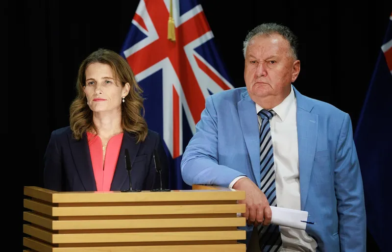 Two people stand at a podium with microphones, in front of New Zealand flags. The woman on the left has wavy brown hair and wears a navy blazer. The man on the right wears a light blue suit and holds papers.