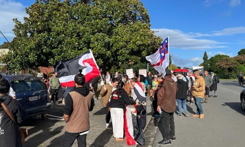 A group of people gather on a street under a large tree, holding flags and signs. Some wear traditional clothing, while others are in casual attire. Cars are parked nearby, and the sky is partly cloudy.